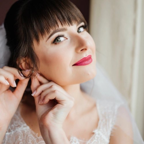 ¿Qué tratamientos faciales hacerte antes de una boda? 1 Novia con maquillaje natural y labios rosados colocándose un pendiente frente a la luz de una ventana.